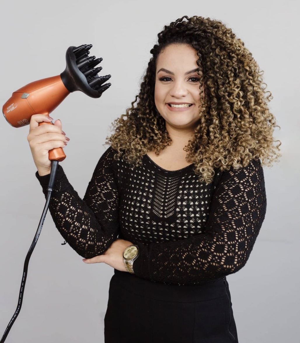 Rebeca Fajardo sorridente com cabelo cacheado e difusor na mão